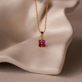 Ombre Ruby Flower Necklace featuring a gold chain and square pink gemstone pendant, displayed on a wavy, cream-colored surface.