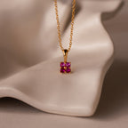 Ombre Ruby Flower Necklace featuring a gold chain and square pink gemstone pendant, displayed on a wavy, cream-colored surface.
