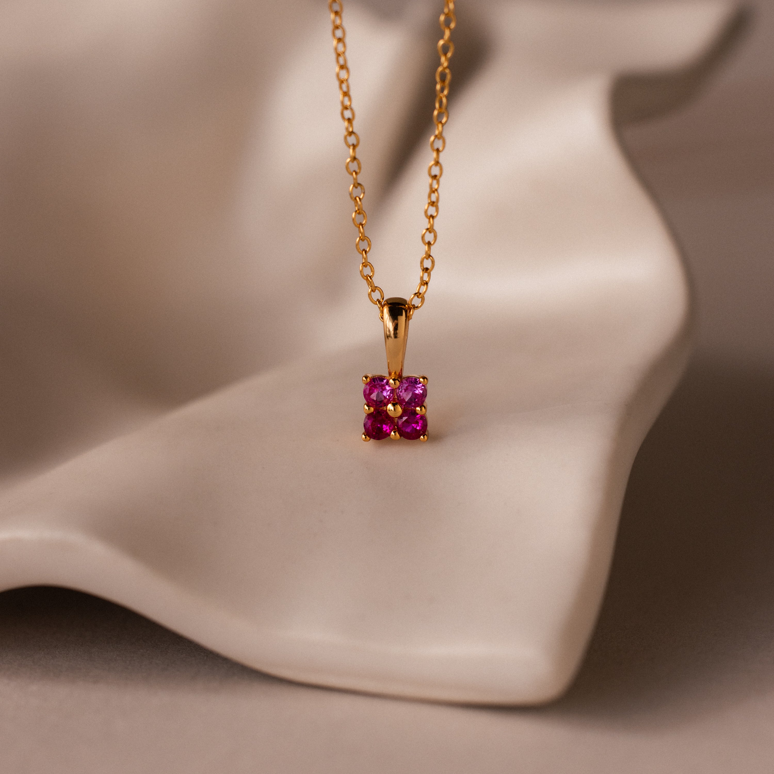 Ombre Ruby Flower Necklace featuring a gold chain and square pink gemstone pendant, displayed on a wavy, cream-colored surface.