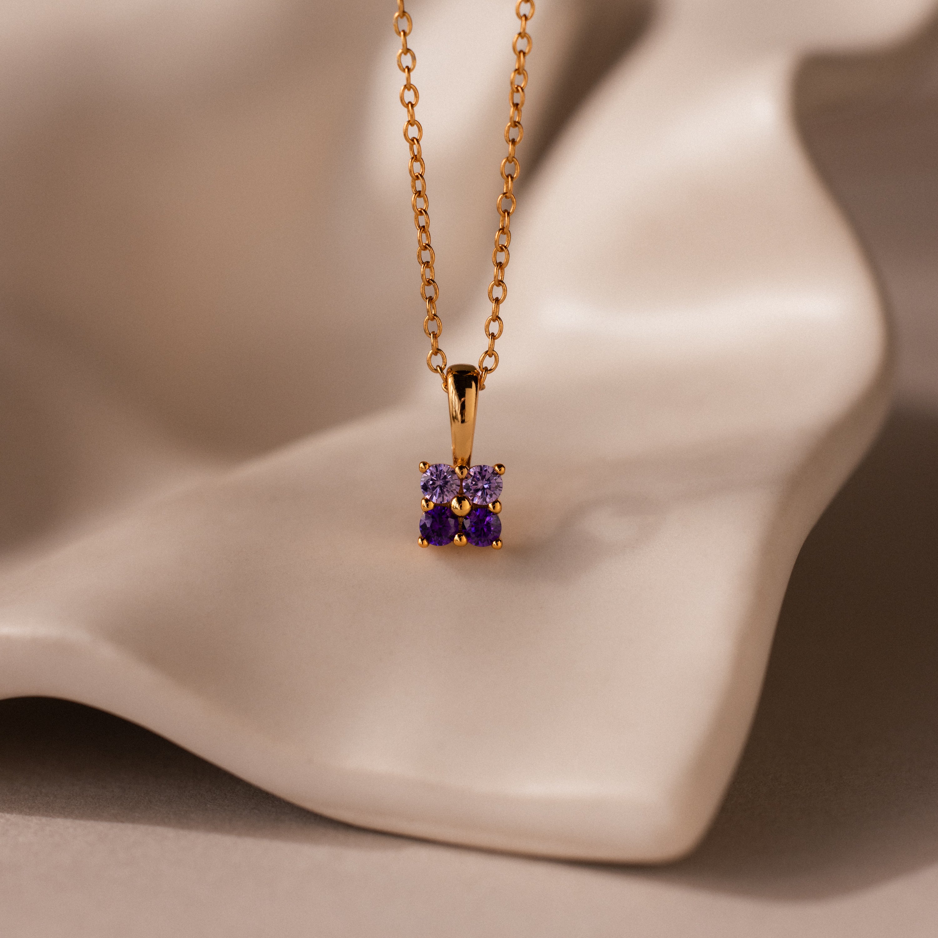 The Ombre Amethyst Flower Necklace features a gold flower pendant adorned with small purple gemstones, displayed on a soft, cream-colored surface.
