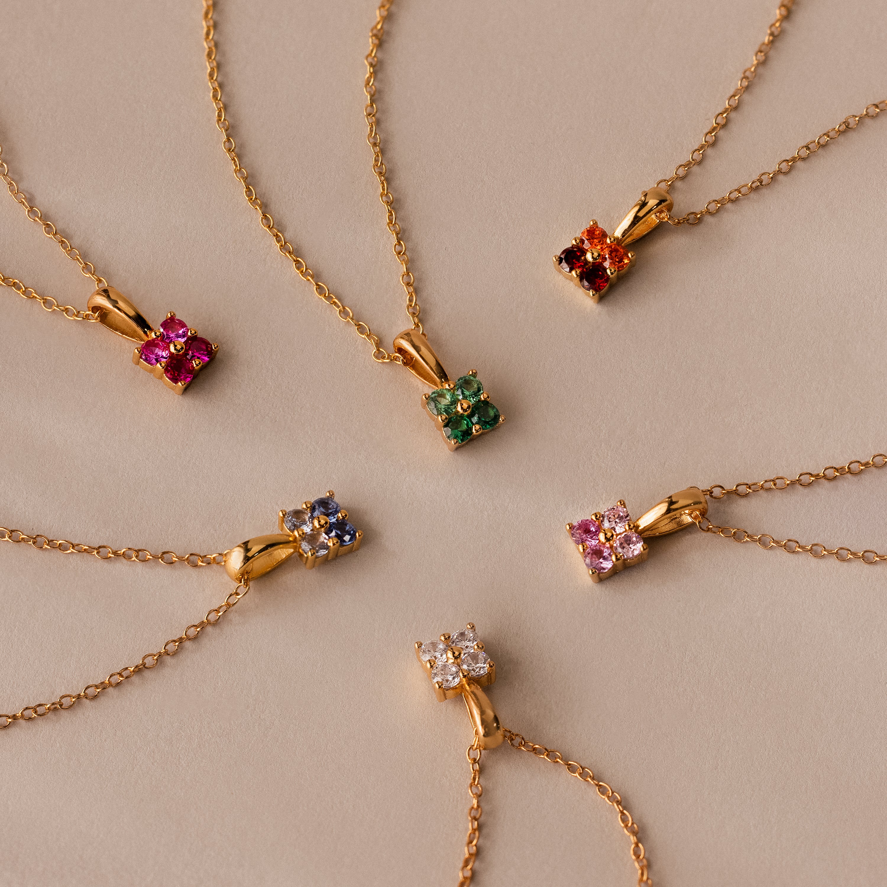 Six gold necklaces with vibrant gemstone pendants, including the Ombre Birthstone Flower Necklace, are elegantly arranged in a circle on a beige background.