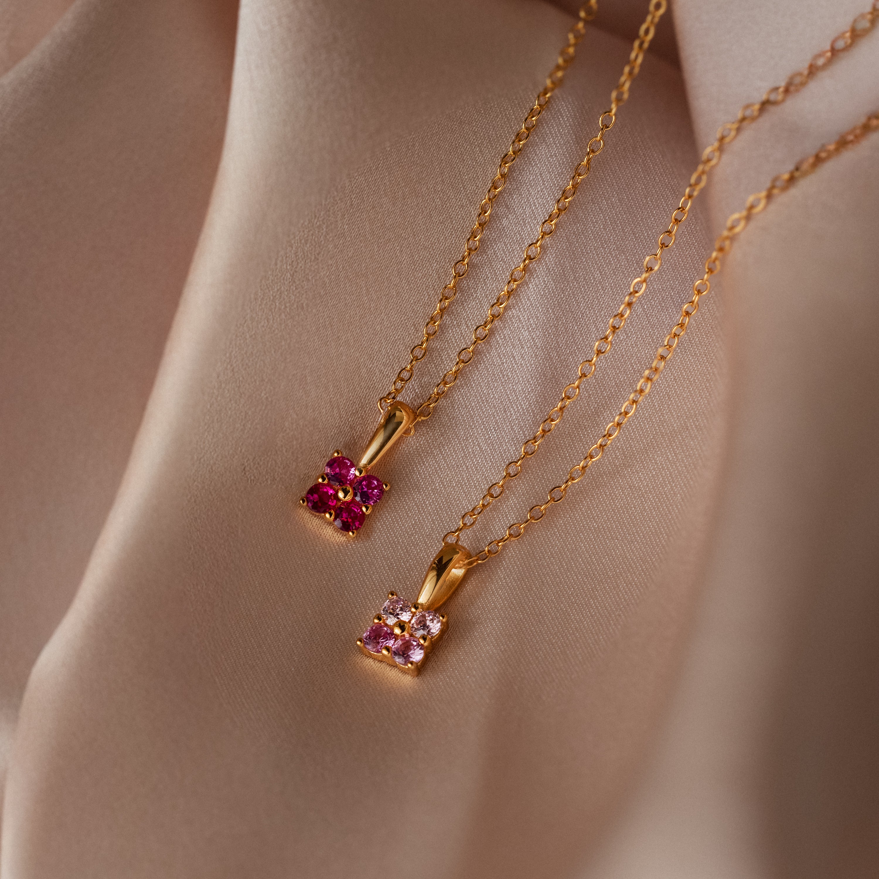 The Ombre Ruby Flower Necklace and another gold piece are displayed on soft, beige satin fabric.