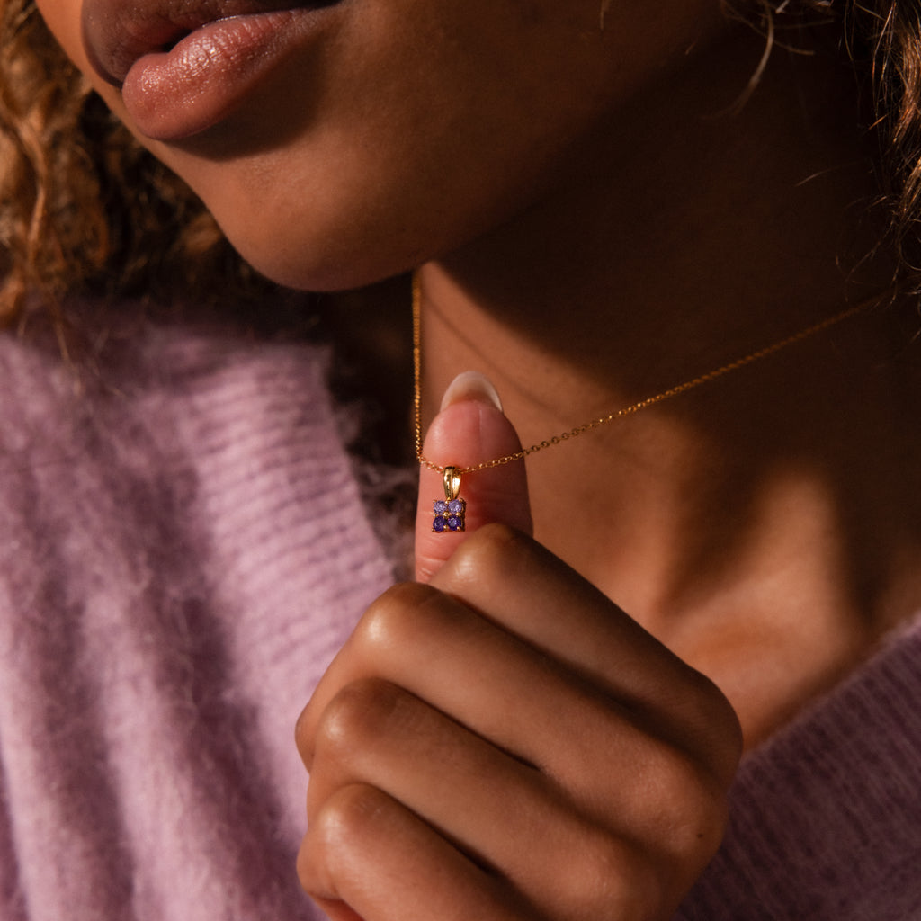 A woman in a purple sweater holds an Ombre Ruby Flower Necklace with a small ruby gemstone pendant near her neck.