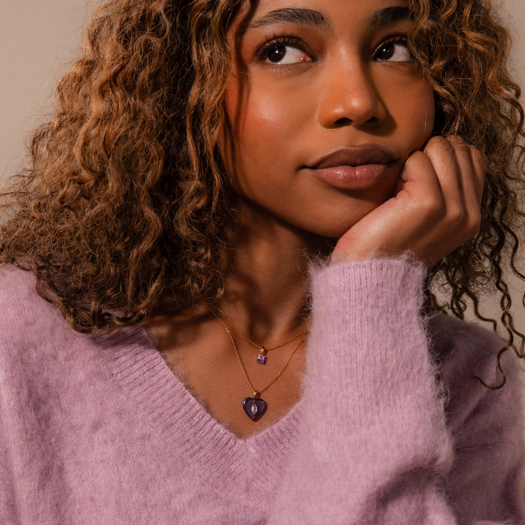 A woman with curly hair in a fuzzy lavender sweater wears a gold Alexandrite Crystal Necklace layered with another gold chain, looking thoughtfully upward.