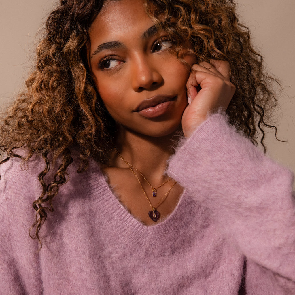 A woman with curly hair in a soft purple sweater gazes thoughtfully to the side, wearing layered necklaces including the Pink Tourmaline Crystal Necklace.
