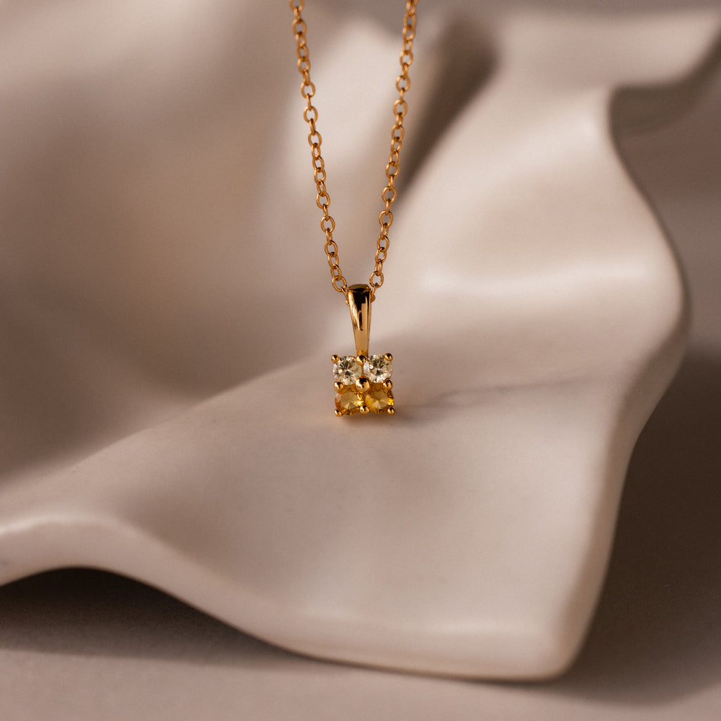 The Ombre Topaz Flower Necklace features a square yellow gemstone pendant and is displayed on a cream-colored, wavy surface.