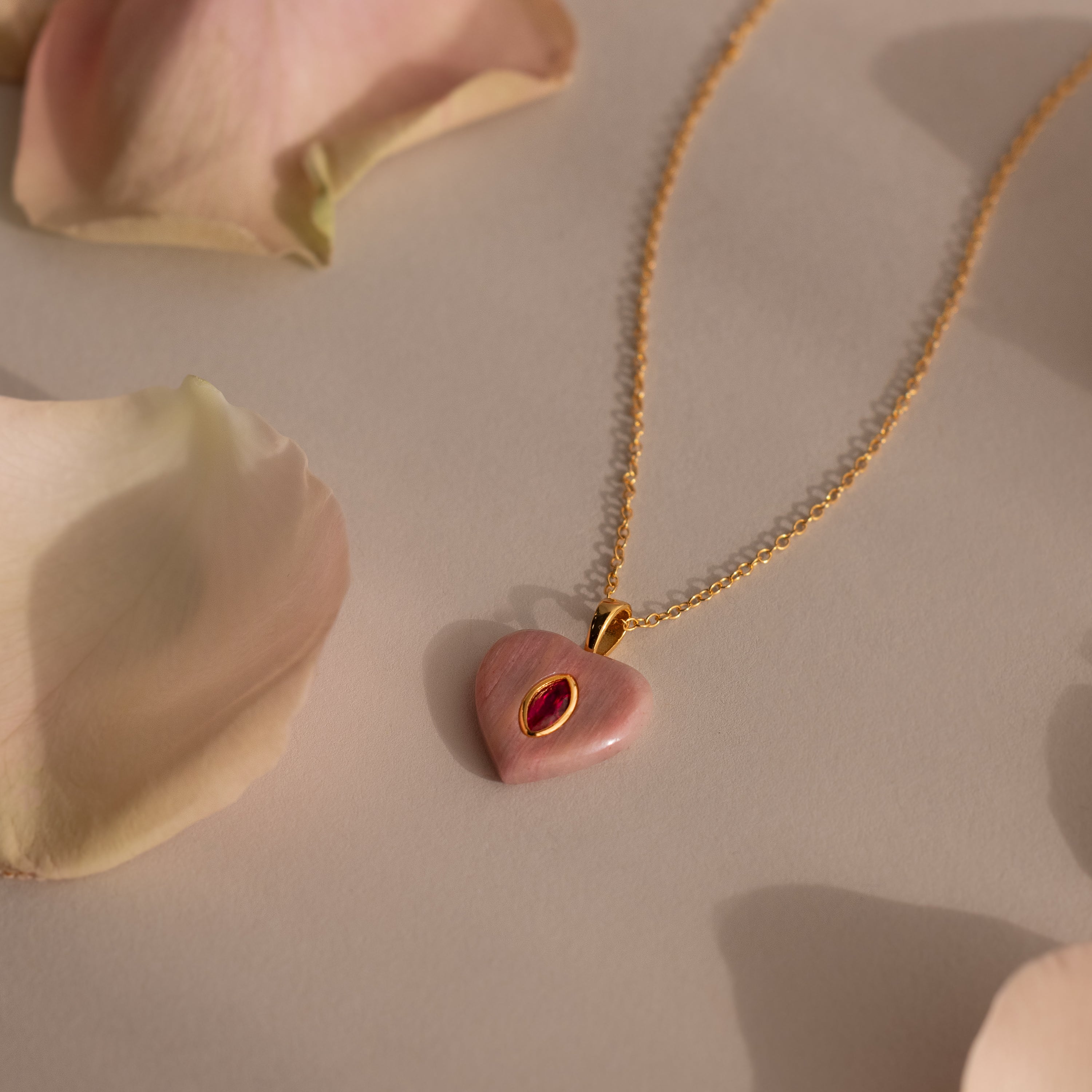 The Ruby Crystal Necklace features a heart-shaped pink jewel, bezel set like a birthstone, and adorned with pale rose petals.