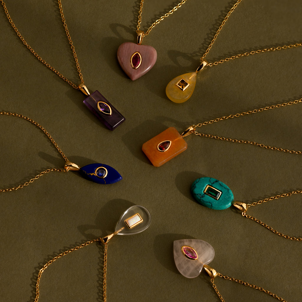 The Pink Tourmaline Crystal Necklace, along with eight other gemstone pendants in bezel settings, hangs on gold chains arranged in a circle against an olive green background.