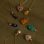 The Pink Tourmaline Crystal Necklace, along with eight other gemstone pendants in bezel settings, hangs on gold chains arranged in a circle against an olive green background.