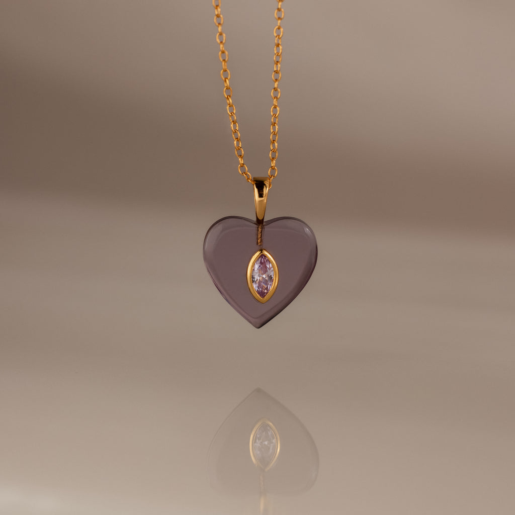 Alexandrite Crystal Necklace: A heart-shaped gold pendant with a central gem, elegantly hanging from a gold chain and showcased on a soft beige background. Perfect as birthstone jewelry or a stunning Birthstone Crystal Necklace for any occasion.
