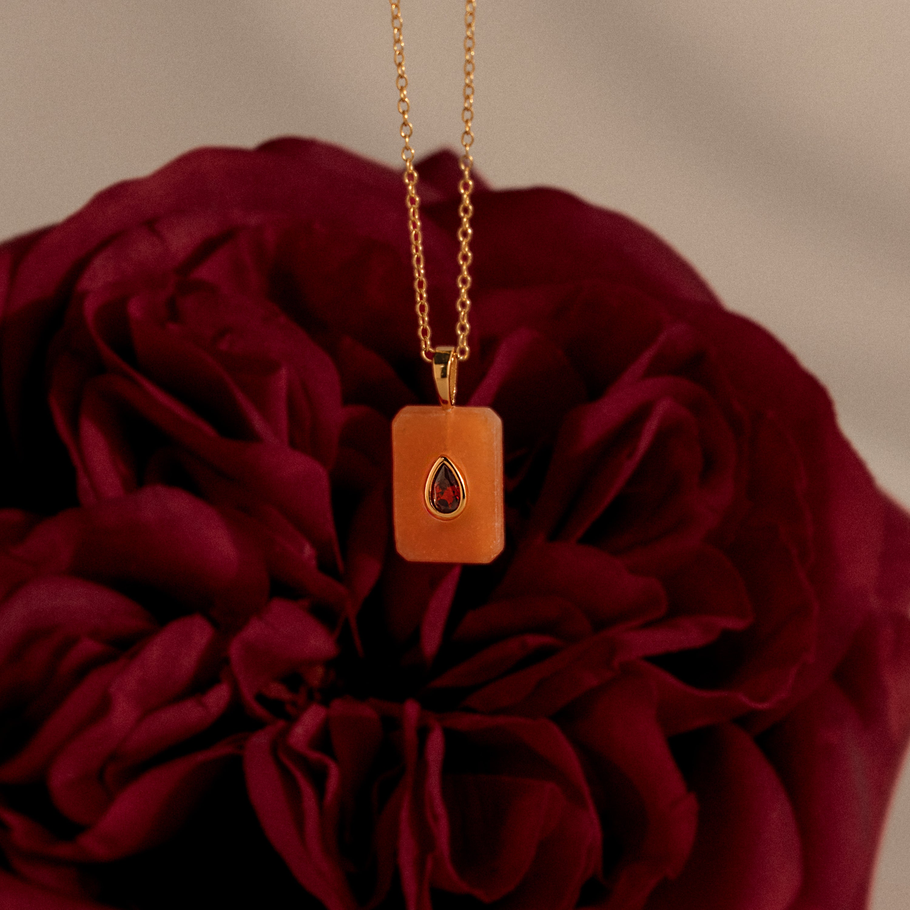 The Garnet Crystal Necklace, highlighted against the rich petals of a dark red flower, is a stunning birthstone jewelry piece with an orange pendant—perfect for any occasion.