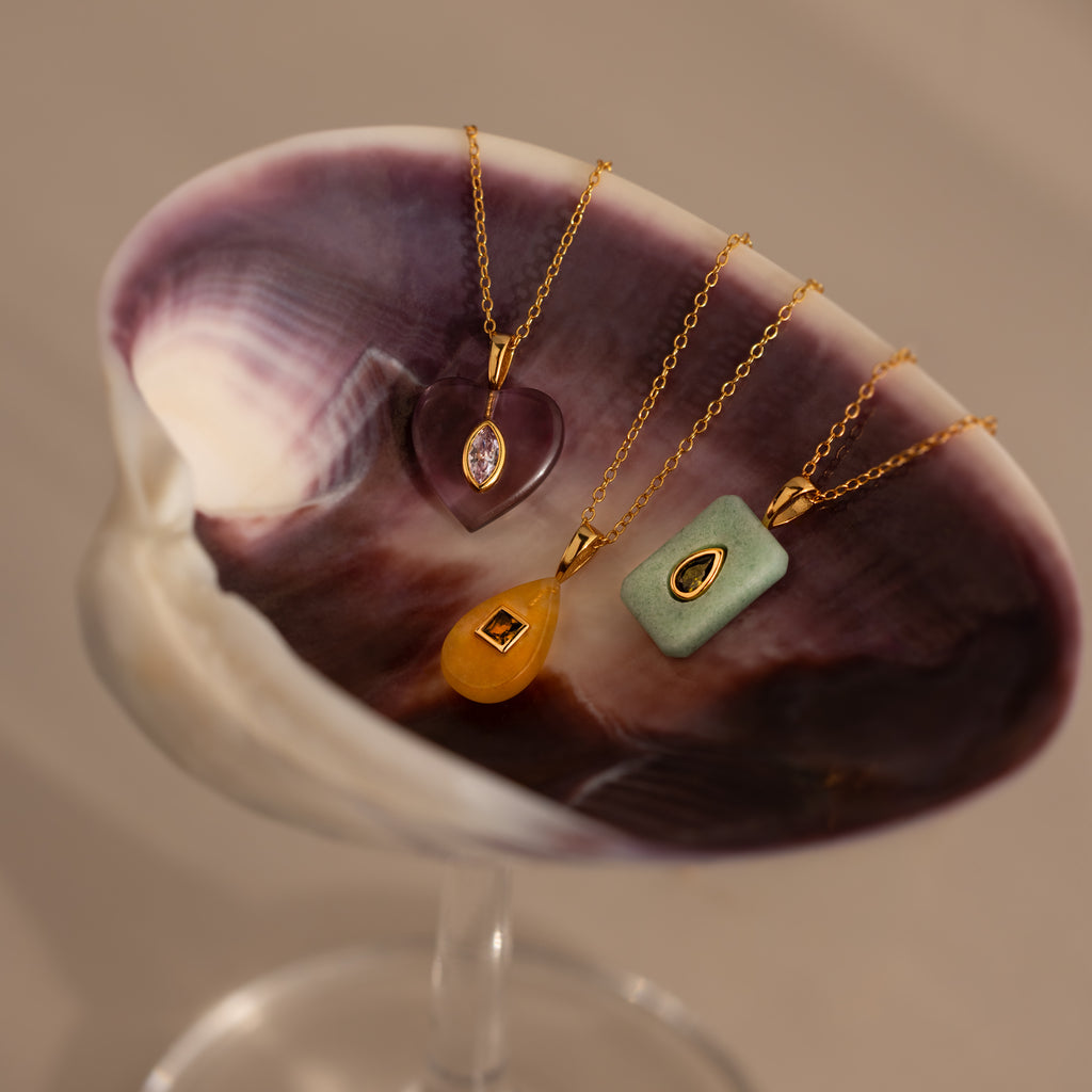Three Birthstone Crystal Necklaces with colorful pendants are displayed on a purple and white seashell.