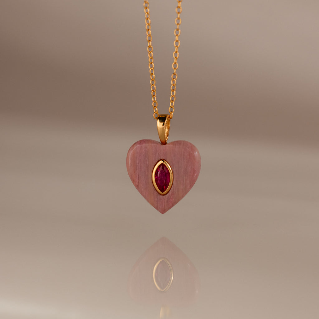 The Ruby Crystal Necklace features a gold chain with a pink heart-shaped pendant, showcasing a bezel-set ruby crystal at its center and accented in gold.