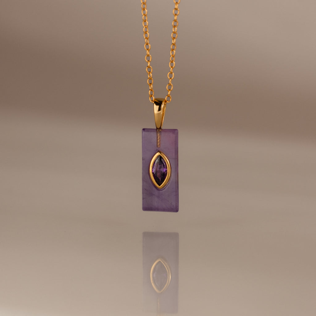 Gold chain necklace with a rectangular purple pendant and gold accent, reflected on a smooth surface—an elegant Amethyst Crystal Necklace perfect for adding a touch of birthstone jewelry to any look.