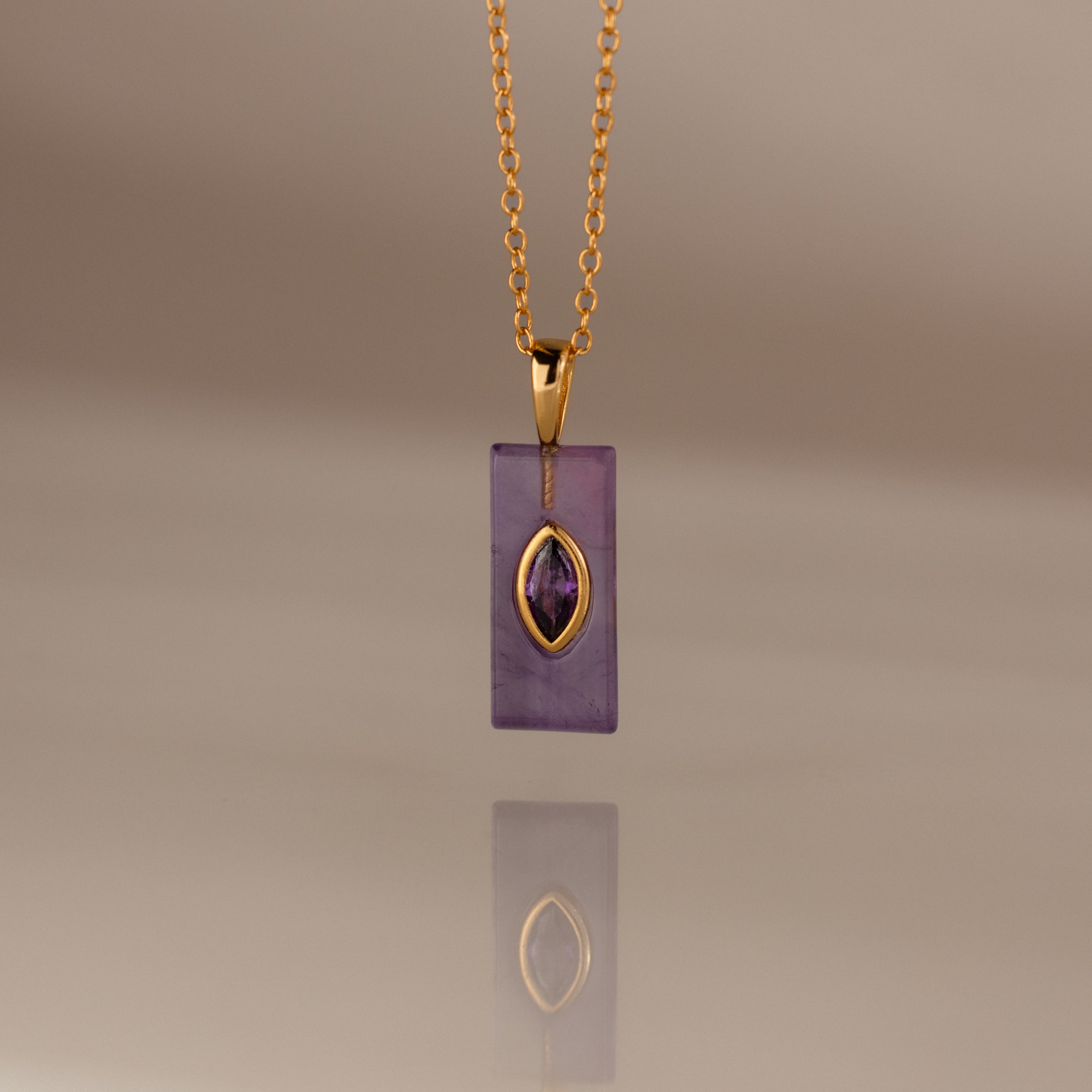 Gold chain necklace with a rectangular purple pendant and gold accent, reflected on a smooth surface—an elegant Amethyst Crystal Necklace perfect for adding a touch of birthstone jewelry to any look.