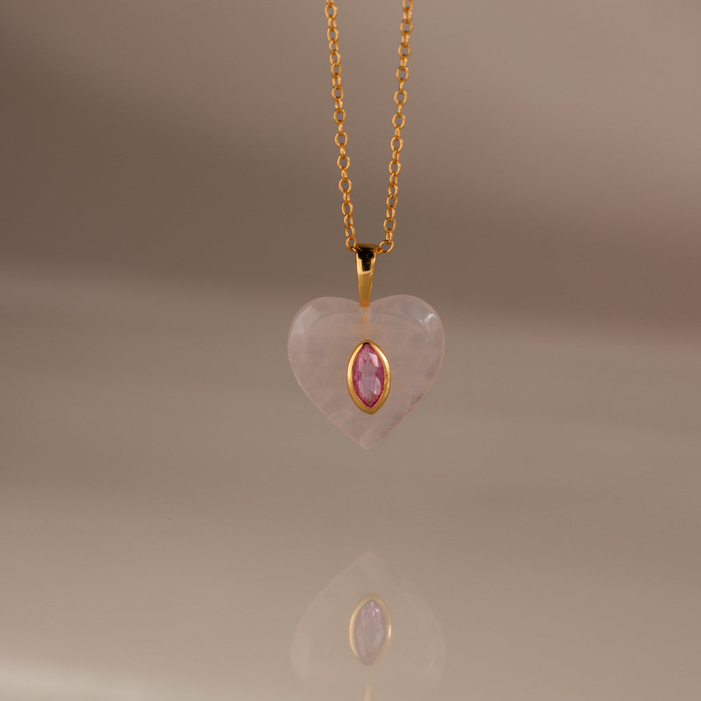 The Pink Tourmaline Crystal Necklace features a gold chain with a heart-shaped pendant, showcasing a bezel-set pink tourmaline birthstone at the center, displayed on a neutral background.