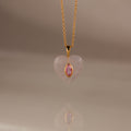 The Pink Tourmaline Crystal Necklace features a gold chain with a heart-shaped pendant, showcasing a bezel-set pink tourmaline birthstone at the center, displayed on a neutral background.