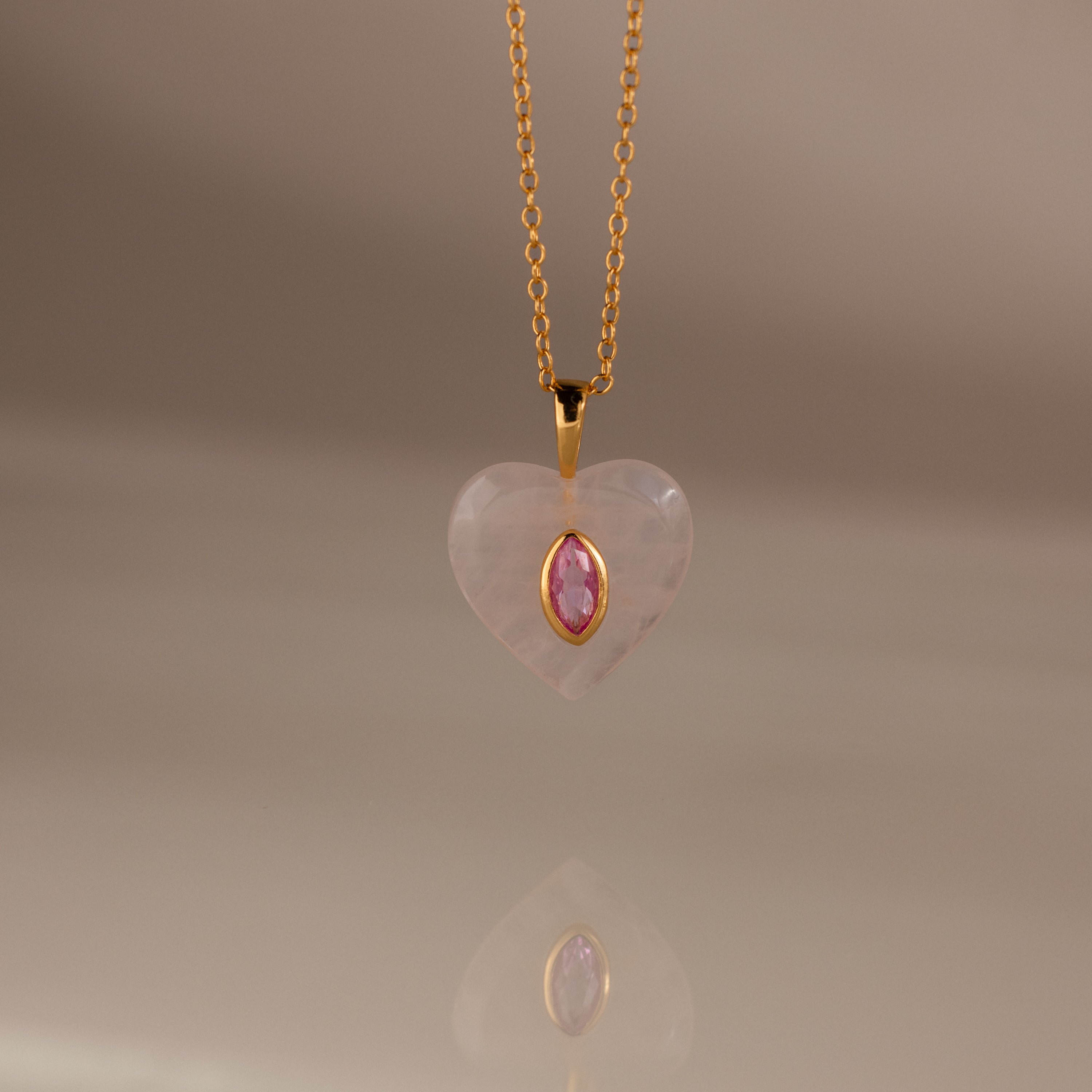 The Pink Tourmaline Crystal Necklace features a gold chain with a heart-shaped pendant, showcasing a bezel-set pink tourmaline birthstone at the center, displayed on a neutral background.