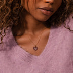 Woman wearing a lilac sweater and the Birthstone Crystal Necklace, featuring a gold chain with a purple heart-shaped birthstone crystal pendant.