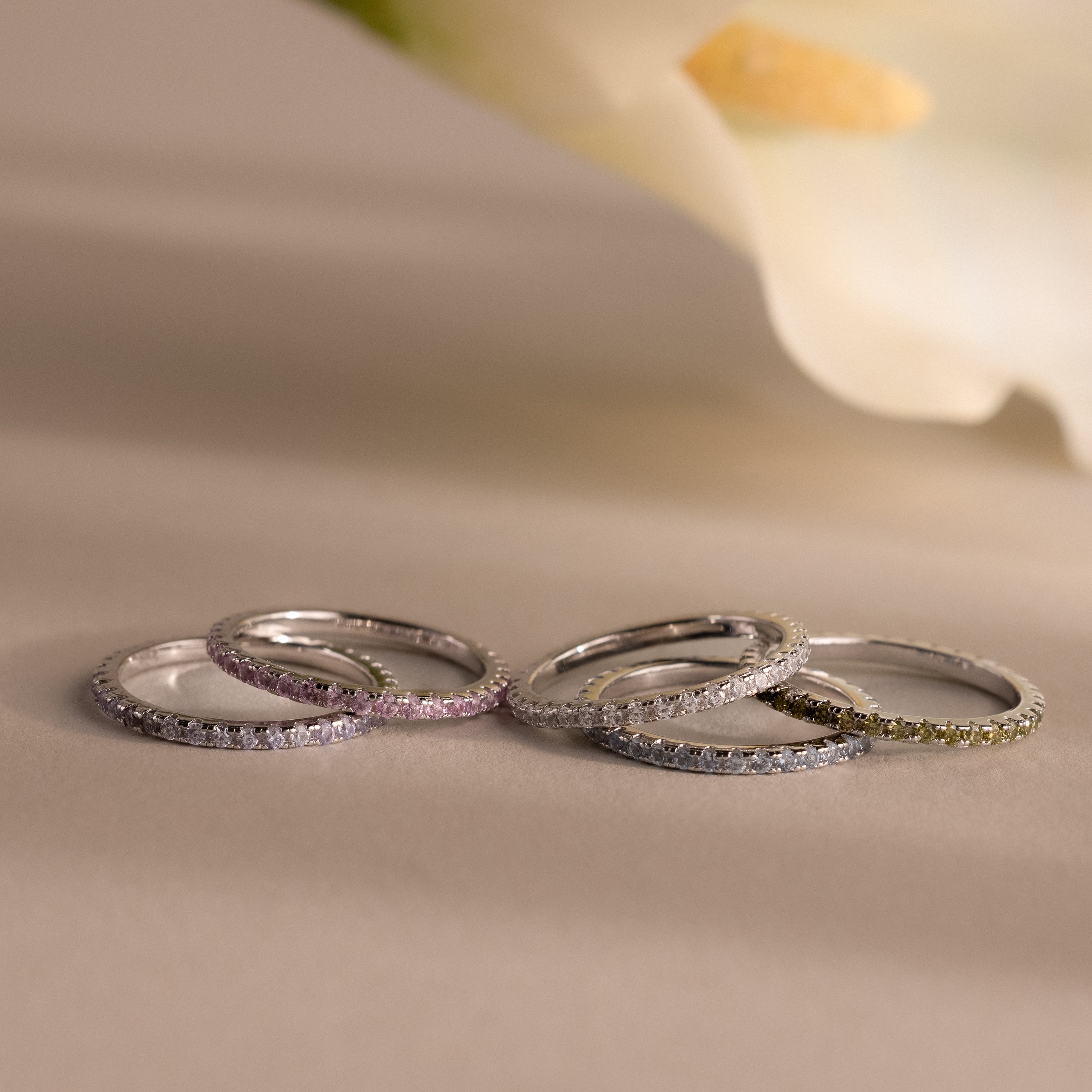 Five Endless Eternity Birthstone Rings, each in a unique pastel shade, are displayed on a neutral surface with a blurred flower in the background, their elegant design adding subtle pops of color to the scene.