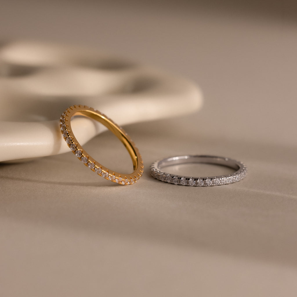 The Endless Eternity Diamond Ring in gold and silver rests on a beige surface, complemented by elegant jewelry and a decorative ceramic piece in the background.