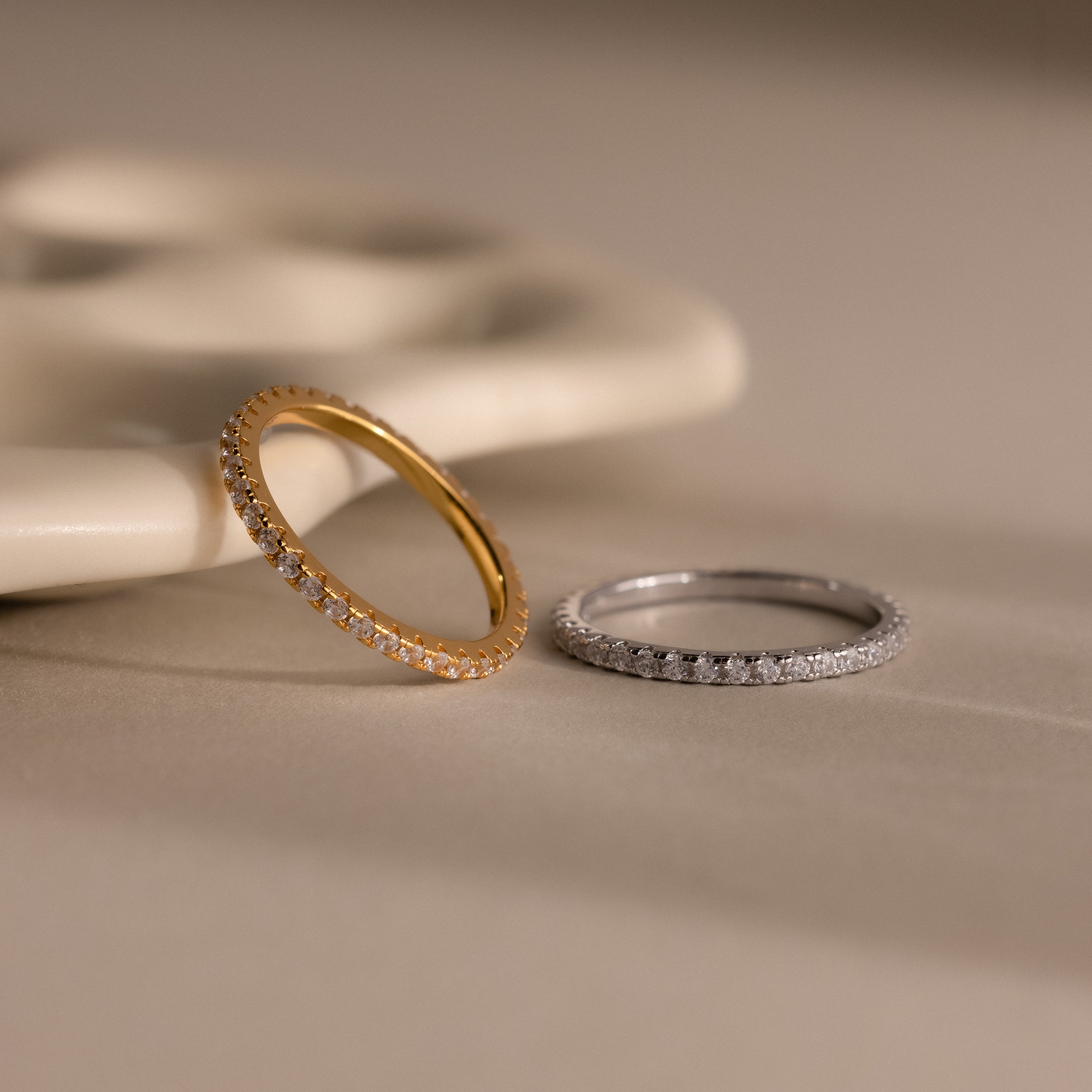 The Endless Eternity Diamond Ring in gold and silver rests on a beige surface, complemented by elegant jewelry and a decorative ceramic piece in the background.
