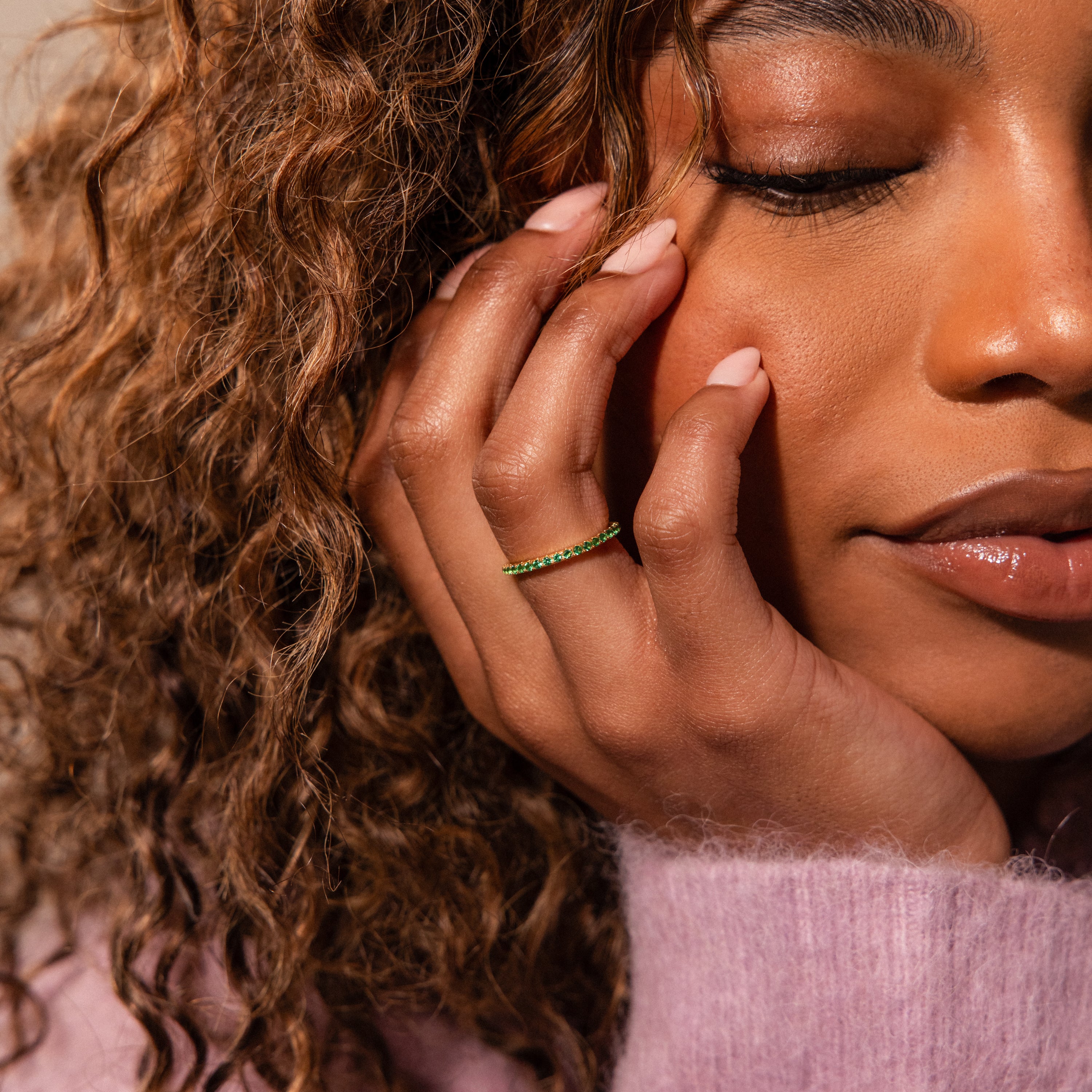 A woman with curly hair touches her face, showcasing an Endless Eternity Blue Zircon Ring while wearing a cozy pink sweater.