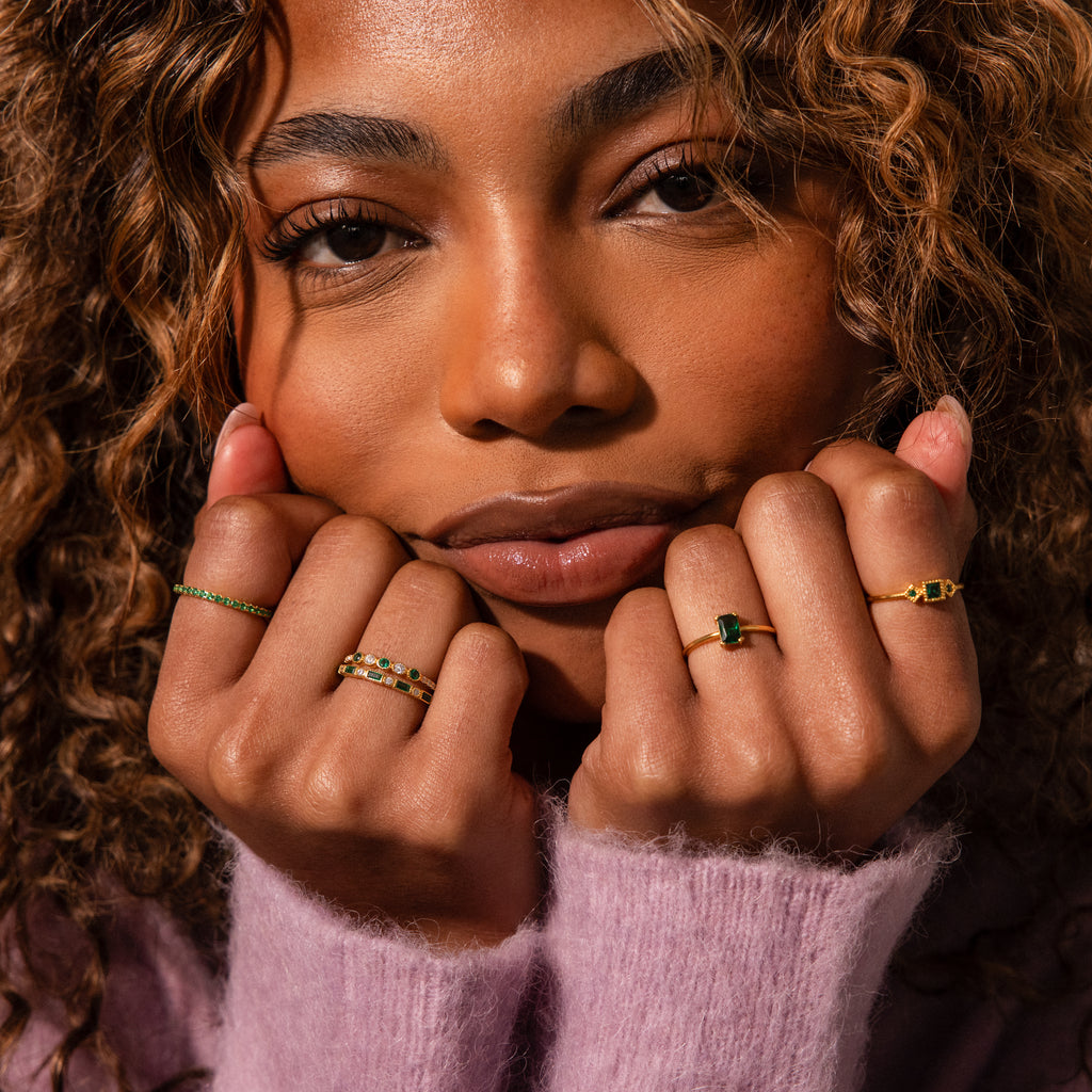 A woman with curly hair, wearing a pink sweater, shows off the Endless Eternity Birthstone Ring featuring sparkling green and white stones, holding her hands near her face.