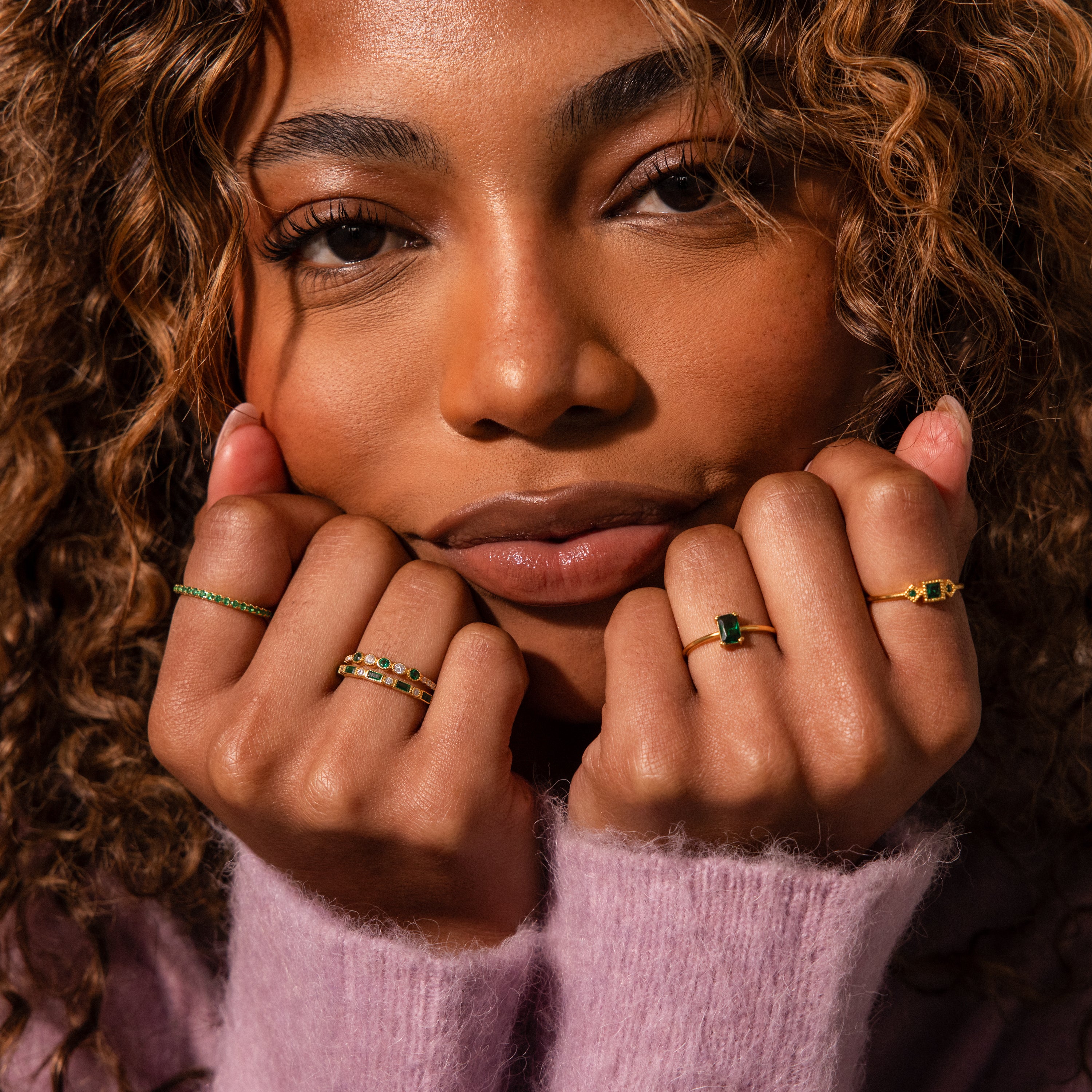 A woman with curly hair, wearing a pink sweater, shows off the Endless Eternity Birthstone Ring featuring sparkling green and white stones, holding her hands near her face.