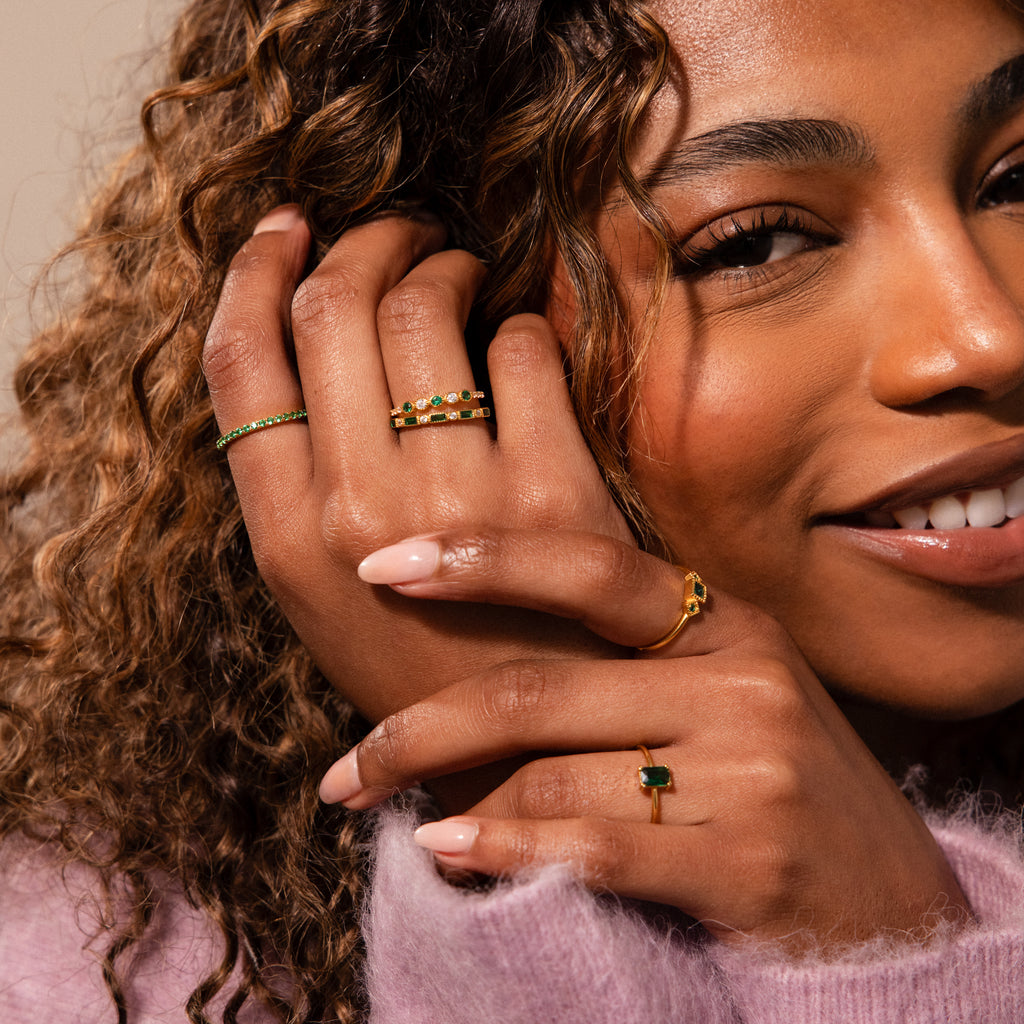 A woman with curly hair smiles, hands adorned with gold rings and an Endless Eternity Pink Tourmaline Ring, paired with striking green gemstone jewelry.