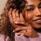 A woman with curly hair smiles, hands adorned with gold rings and an Endless Eternity Pink Tourmaline Ring, paired with striking green gemstone jewelry.