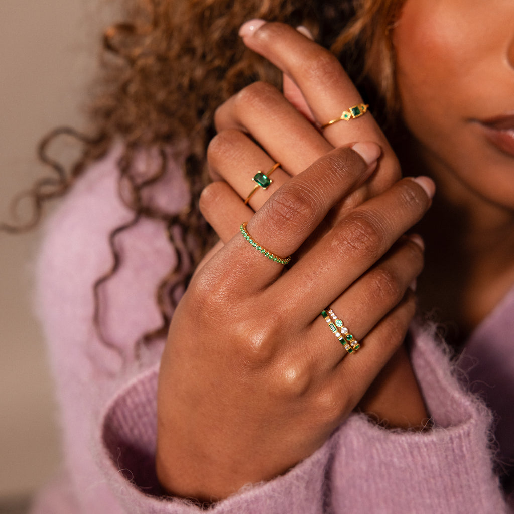 A person with curly hair wears elegant jewelry—multiple gold gemstone rings, including the Endless Eternity Topaz Ring—hands gently crossed in a pink sweater.
