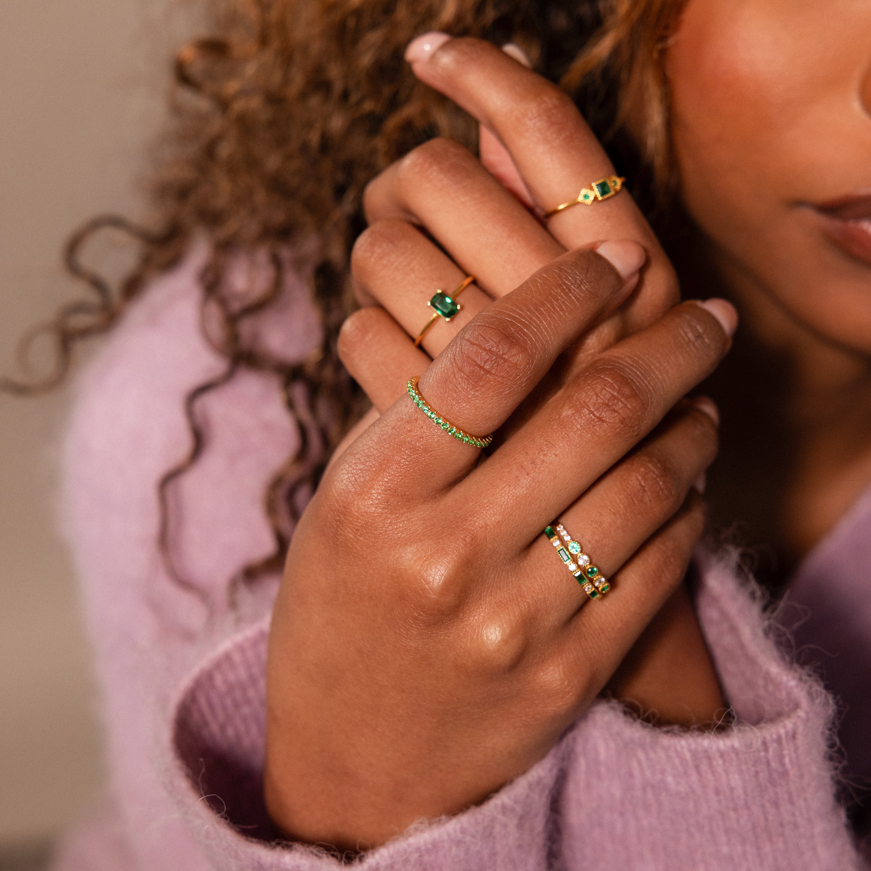 A person with curly hair wears elegant jewelry—multiple gold gemstone rings, including the Endless Eternity Topaz Ring—hands gently crossed in a pink sweater.