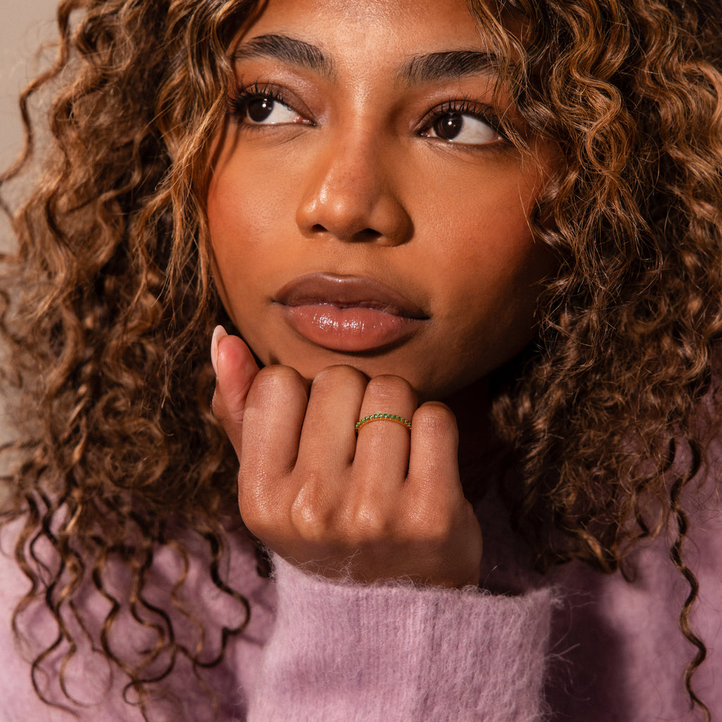 A woman with curly hair in a purple sweater with lace accents rests her chin on her hand, displaying the Lace Eternity Garnet Ring.