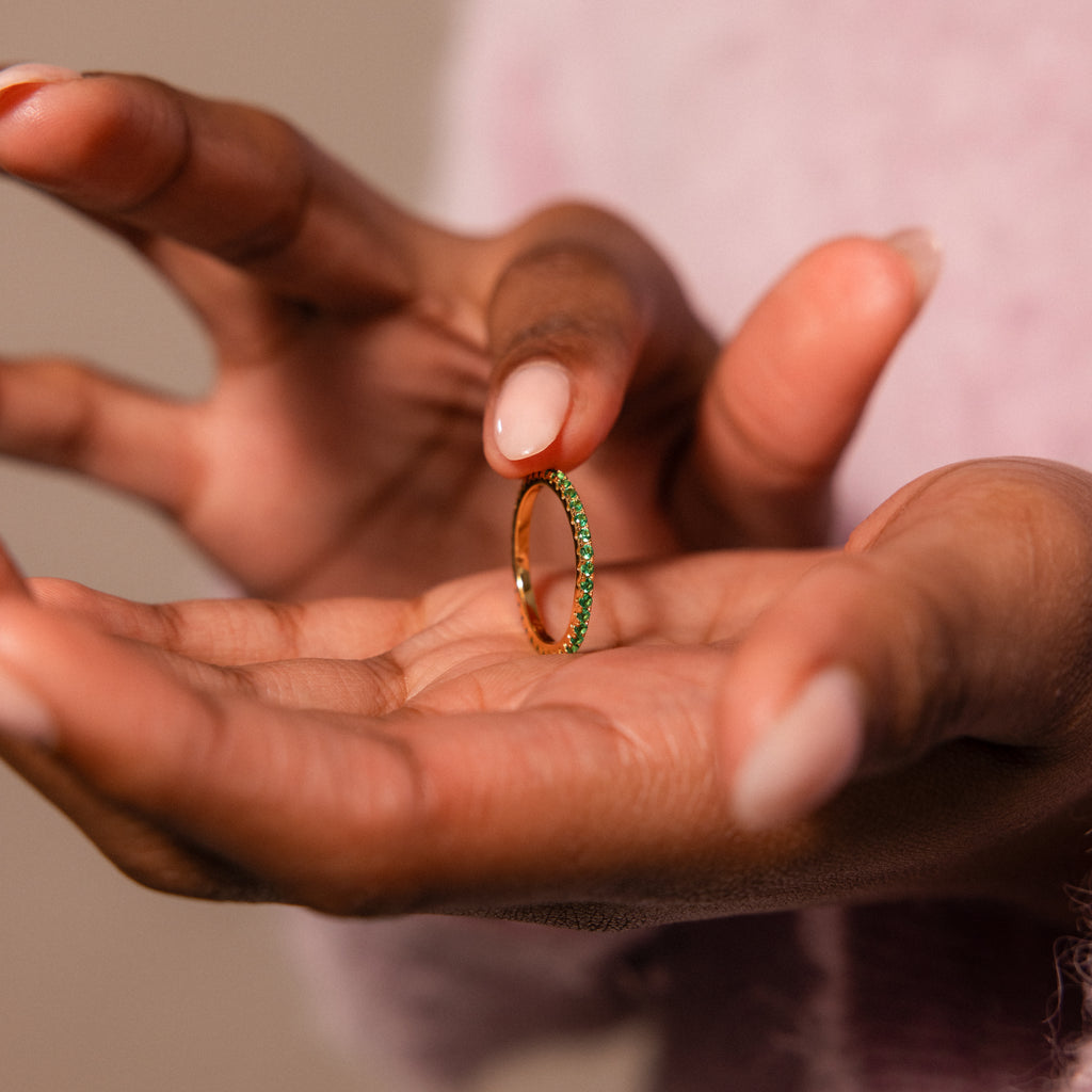 A person holds an elegant Endless Eternity Topaz Ring featuring green stones between their thumb and forefinger.