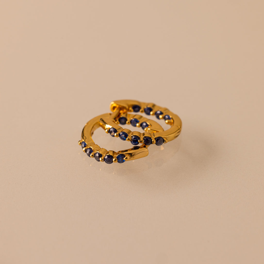 Two gold Inside Out Sapphire Huggies, featuring small dark blue gemstones ideal for stacking, are displayed on a beige surface.