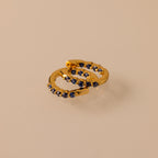 Two gold Inside Out Sapphire Huggies, featuring small dark blue gemstones ideal for stacking, are displayed on a beige surface.