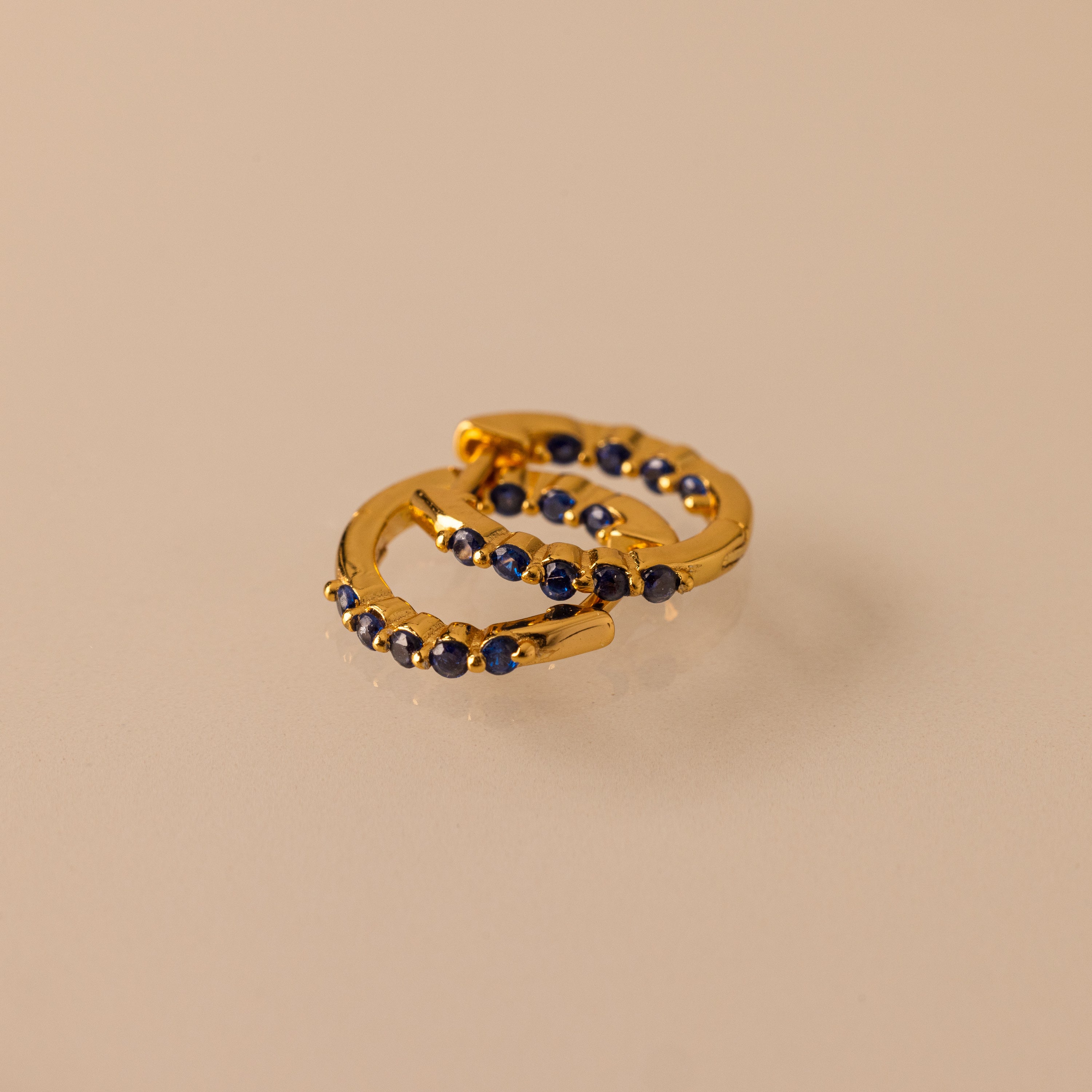 Two gold Inside Out Sapphire Huggies, featuring small dark blue gemstones ideal for stacking, are displayed on a beige surface.