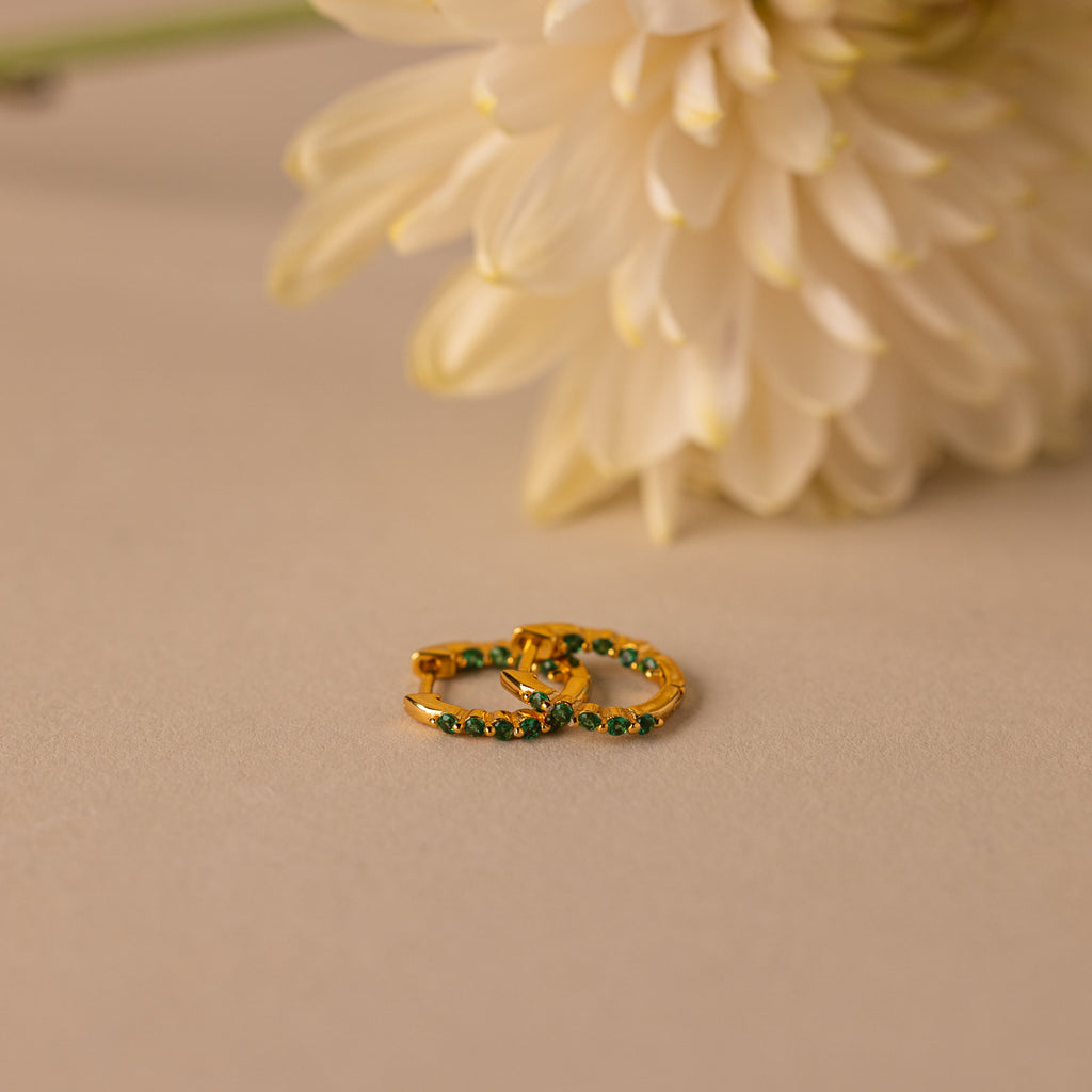 Inside Out Emerald Huggies, gold hoop earrings with green stones, are perfect Birthstone Huggie Earrings and are displayed on a beige surface with a white flower in the background.