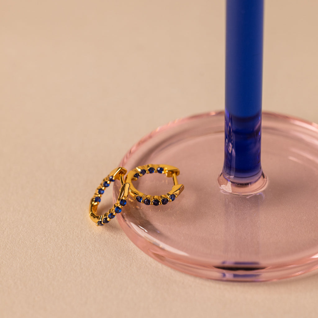 The Inside Out Sapphire Huggies, featuring blue gemstones, are displayed beside a pink and blue glass candlestick holder—ideal for creating stylish stacked earring looks.