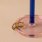 The Inside Out Sapphire Huggies, featuring blue gemstones, are displayed beside a pink and blue glass candlestick holder—ideal for creating stylish stacked earring looks.