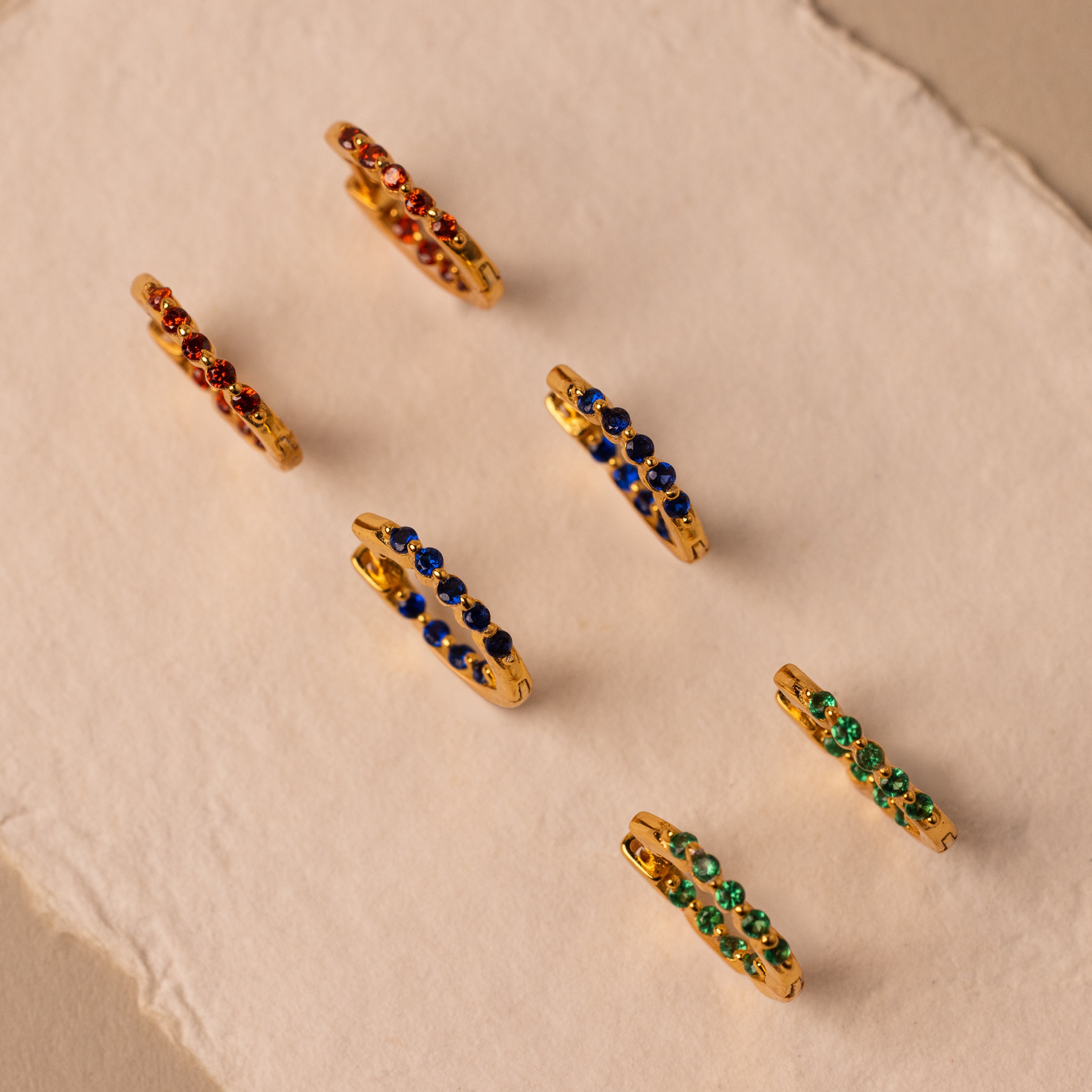 Six Inside Out Sapphire Huggies with red, blue, and green gemstones are arranged on textured beige and cream surfaces, perfect for adding a playful gemstone earring pop to any look.