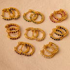 Nine pairs of gold hoop stacking earrings, including Inside Out Ruby Huggies with colorful gemstone accents, are arranged on a light beige surface.
