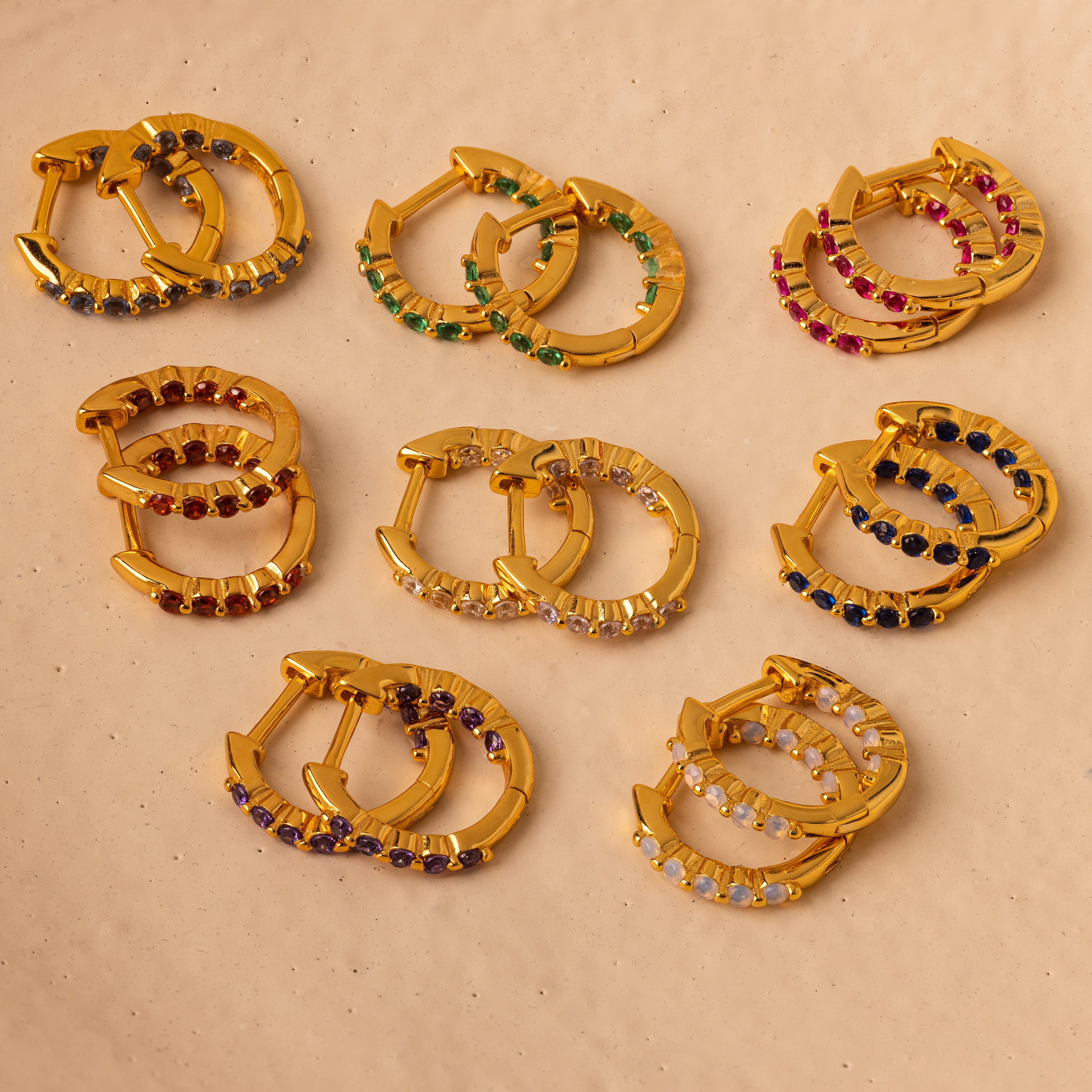 Nine pairs of gold hoop stacking earrings, including Inside Out Ruby Huggies with colorful gemstone accents, are arranged on a light beige surface.
