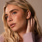 A woman with long blonde hair touches her ear, showing off Inside Out Ruby Huggies while wearing a light purple sweater and gazing off to the side.
