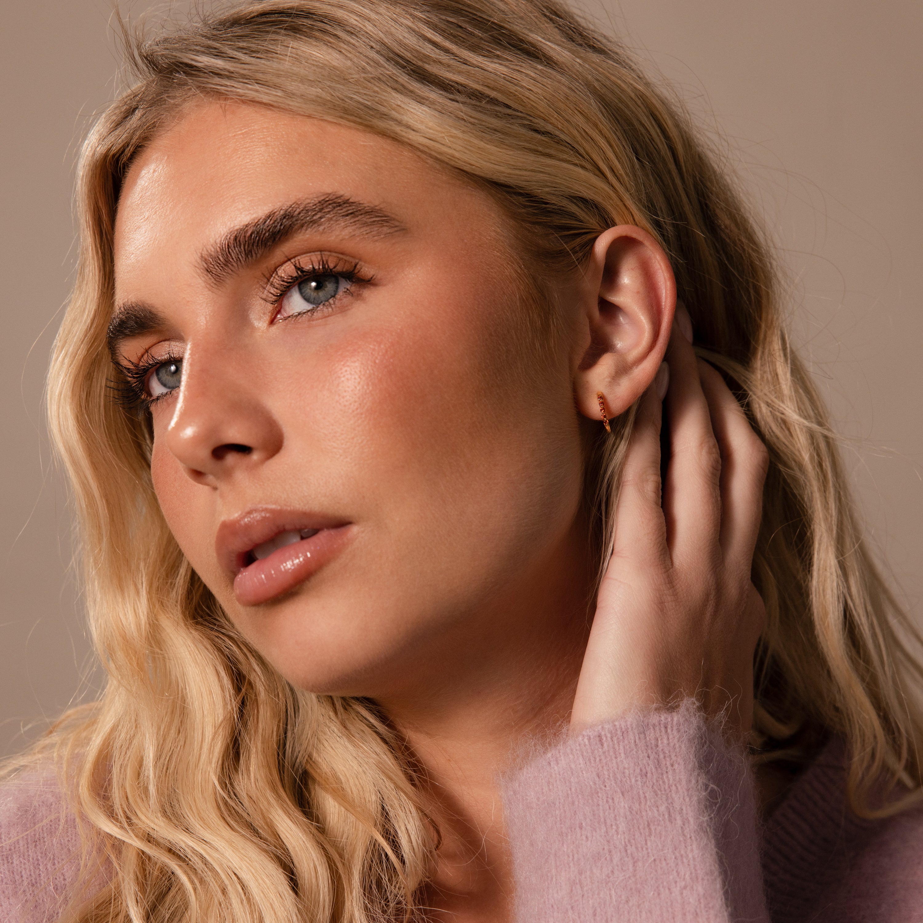 A woman with long blonde hair touches her ear, showing off Inside Out Ruby Huggies while wearing a light purple sweater and gazing off to the side.