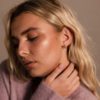 Woman with closed eyes and blonde hair touches her neck, wearing a soft purple sweater and stacking earrings that accent her Inside Out Emerald Huggies.
