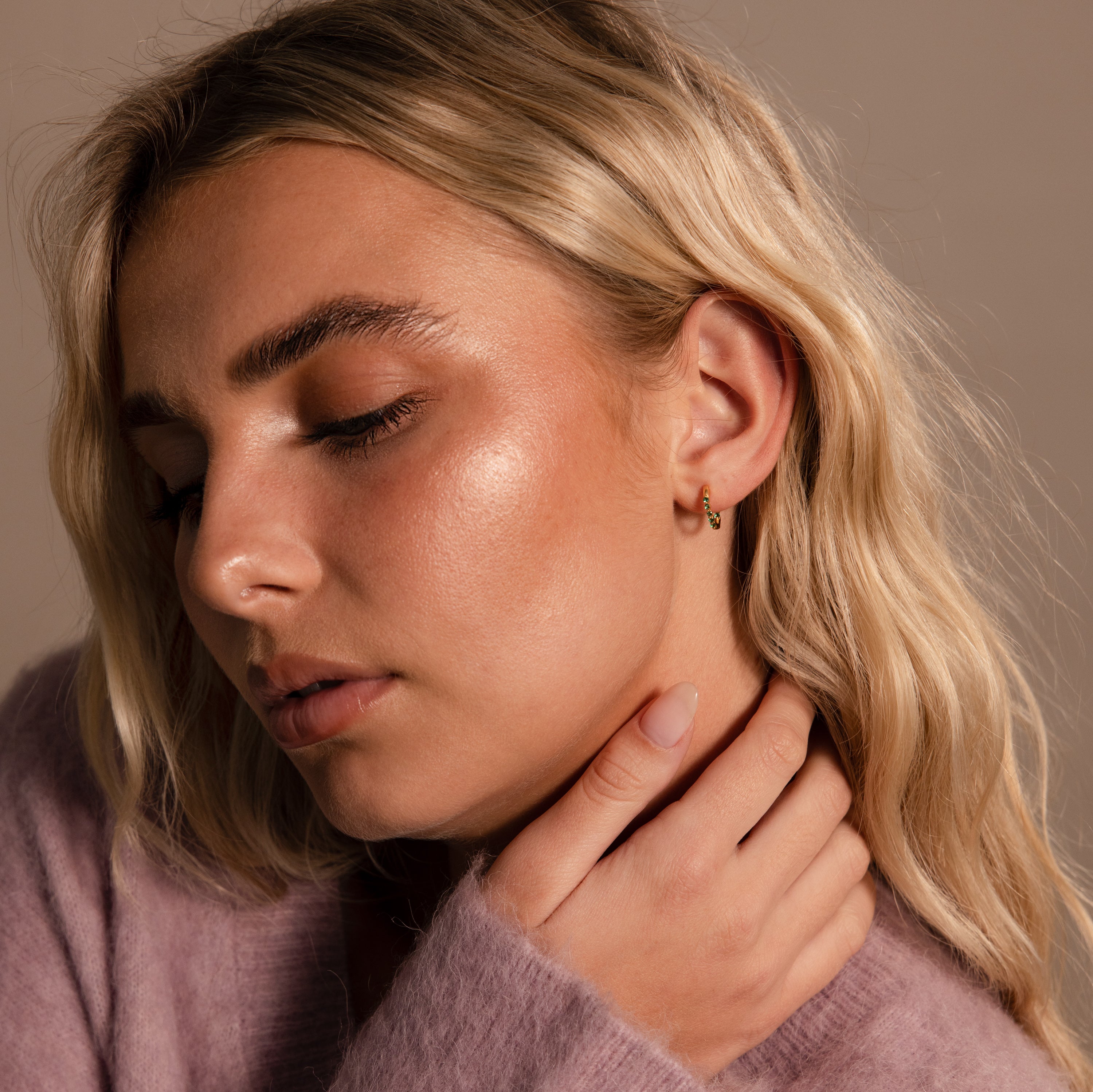 Woman with closed eyes and blonde hair touches her neck, wearing a soft purple sweater and stacking earrings that accent her Inside Out Emerald Huggies.