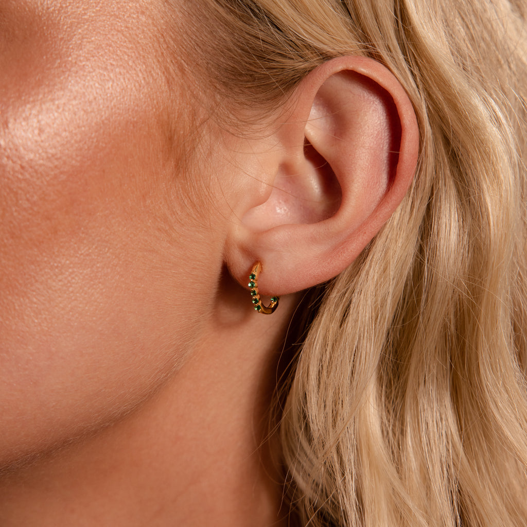 Close-up of a woman's ear wearing Inside Out Sapphire Huggies—small gold earrings set with green stones, ideal for stacking, shown with blonde hair.