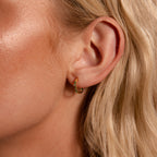 Close-up of a woman's ear wearing Inside Out Emerald Huggies, small gold hoop earrings with green gemstones, ideal for stacking or as chic Birthstone Huggie Earrings, elegantly complemented by her blonde hair.